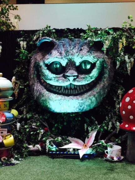 Artificial wisteria trees and ivy for hire around a glowing Cheshire Cat face, red toadstools and stacked teacups