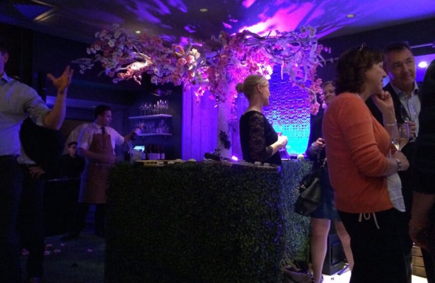 Artificial blossom branches for hire, over a hedge bar uplit purple, with guests and a bartender
