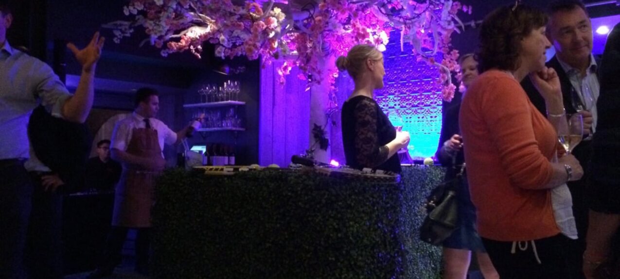 Artificial hedge-fronted bar for hire, topped with pink and white blossom branches and purple lighting