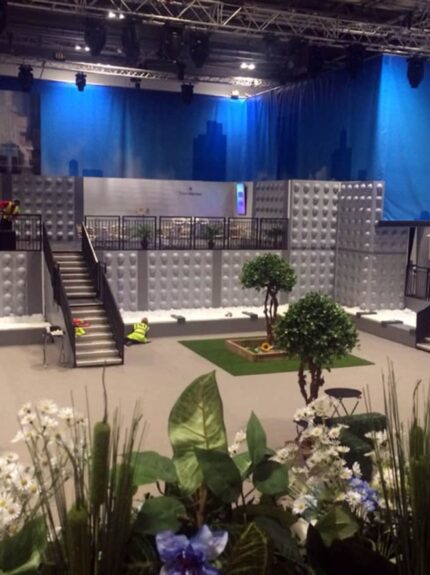 Artificial topiary trees and flower beds for hire in an exhibition hall, with a stepped stage and blue drapes behind