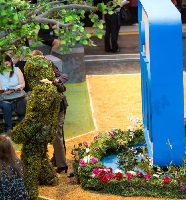 Artificial leafy tree for hire beside a large LinkedIn logo, a flower bed and a moss-covered figure, guests seated nearby