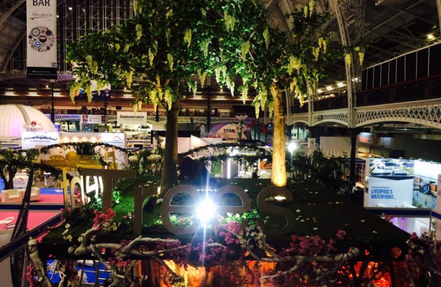 Artificial white wisteria trees for hire, uplit above a large Event Trees sign, beside a Confex Bar banner