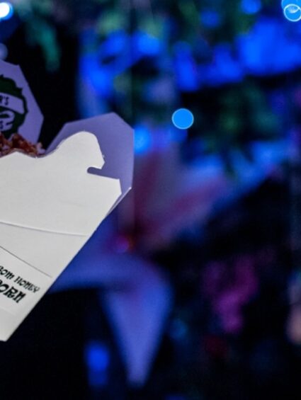 Hanging takeaway popcorn box among artificial foliage and blue fairy lights, close up
