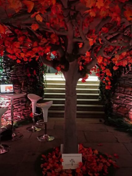 Artificial red tree for hire, with autumn red leaves, beside uplit stone walls, steps and bar stools
