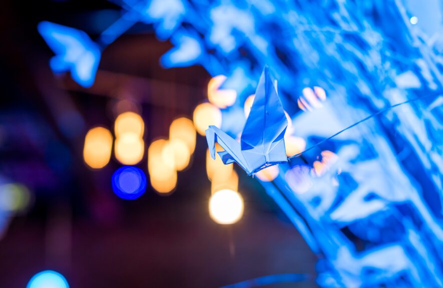 Close-up of a blue-lit paper origami crane hanging on a white branch centrepiece, with golden lights blurred behind