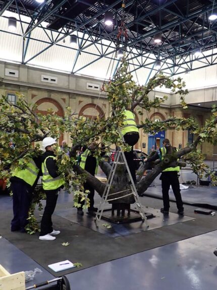 Large bespoke artificial tree for hire, half dressed with leaves, being assembled by crew in hi-vis jackets in a large hall
