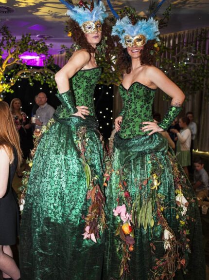 Two stilt walkers in green dresses, feathered masks and woodland costume, with leafy branches behind