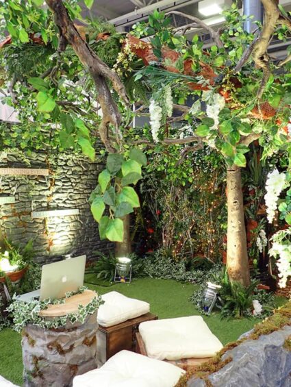 Artificial trees with white wisteria for hire, in a woodland stand with a stone-effect wall, grass and cushion seats