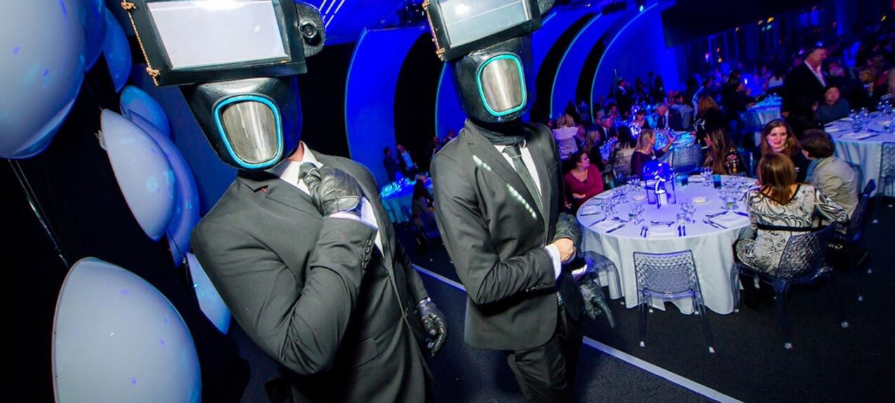 Two performers in retro robot-style head costumes with screens for faces, beside diners in a blue-lit hall