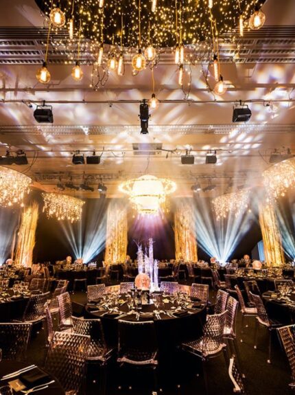 Tall golden twig columns and hanging fairy light chandeliers around a dark banquet hall with spotlights