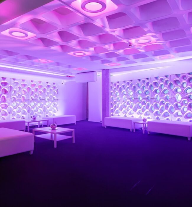 Purple-lit lounge with white sofas, low tables and patterned wall panels