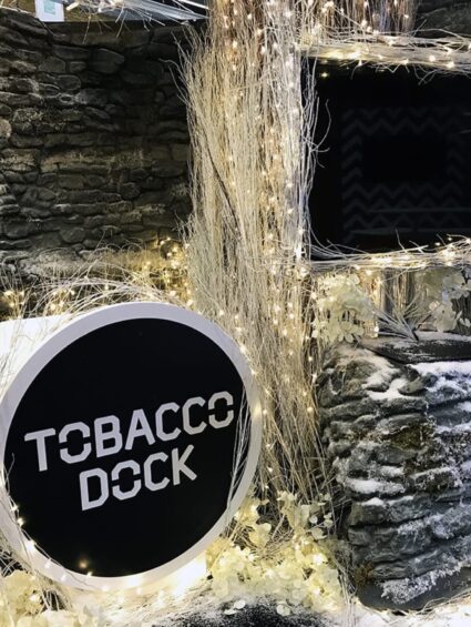 Artificial white twigs with fairy lights for hire, around a stone-effect wall and a round 'TOBACCO DOCK' sign with faux snow