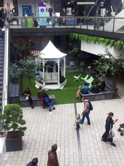 Artificial trees on artificial grass for hire, around a white bandstand and deckchairs in a shopping centre atrium