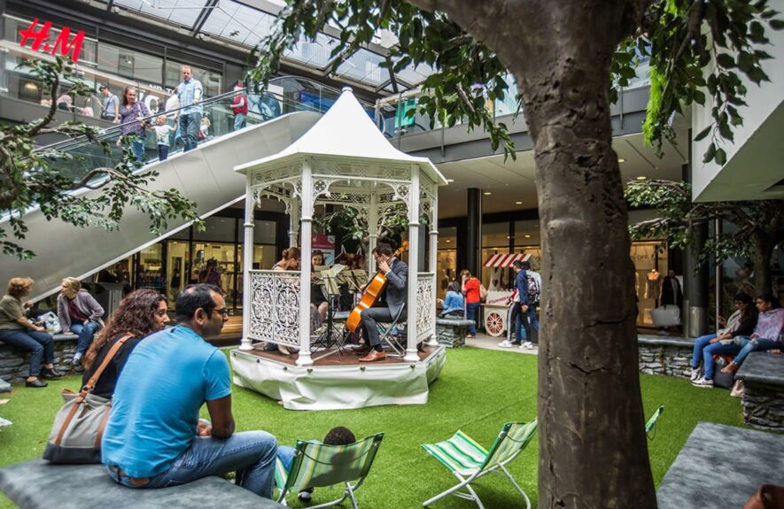 Artificial tree for hire, beside a white bandstand with a cellist on artificial grass near an escalator