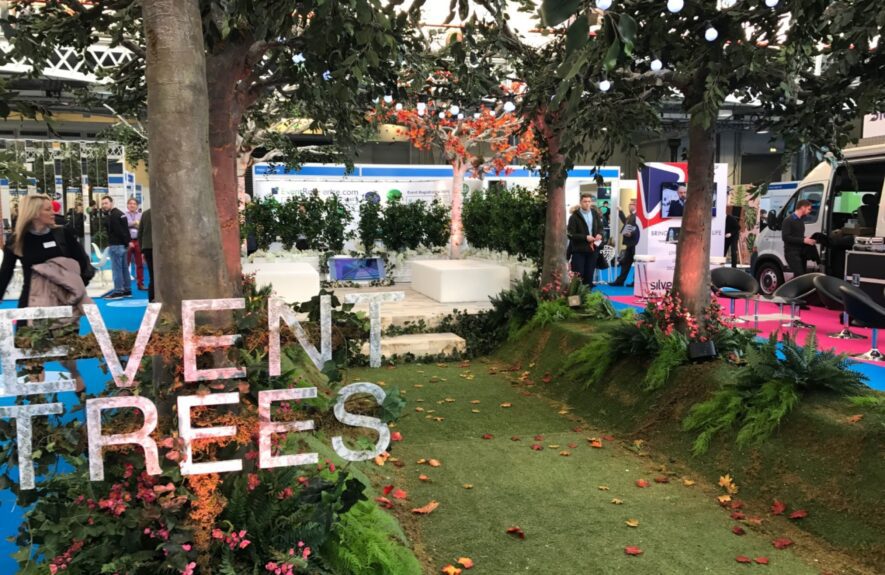 Artificial trees for hire on a grass mound with foliage, autumn leaves and large 'EVENT TREES' letters at an exhibition