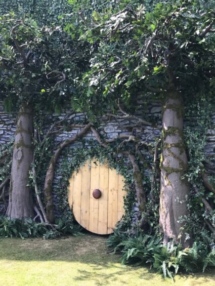 Artificial trees and ivy for hire, built against a stone-effect wall around a round wooden door on a lawn