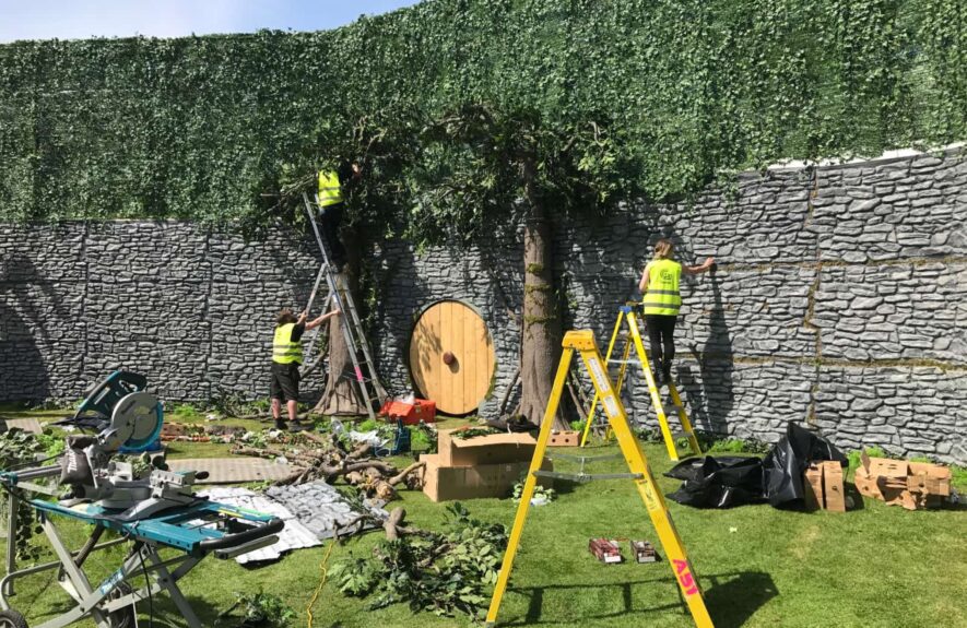Crew installing artificial trees and an ivy wall against a stone-effect wall, with ladders and a round wooden door