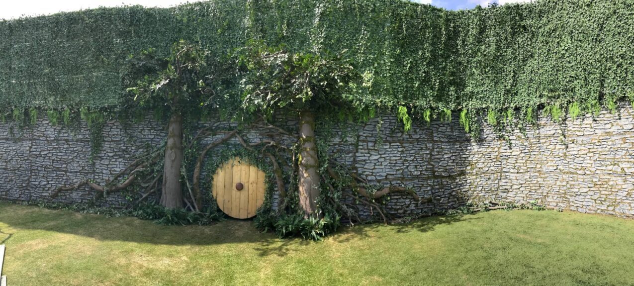Wide view of artificial trees and an ivy wall for hire, built against a stone-effect wall with a round wooden door