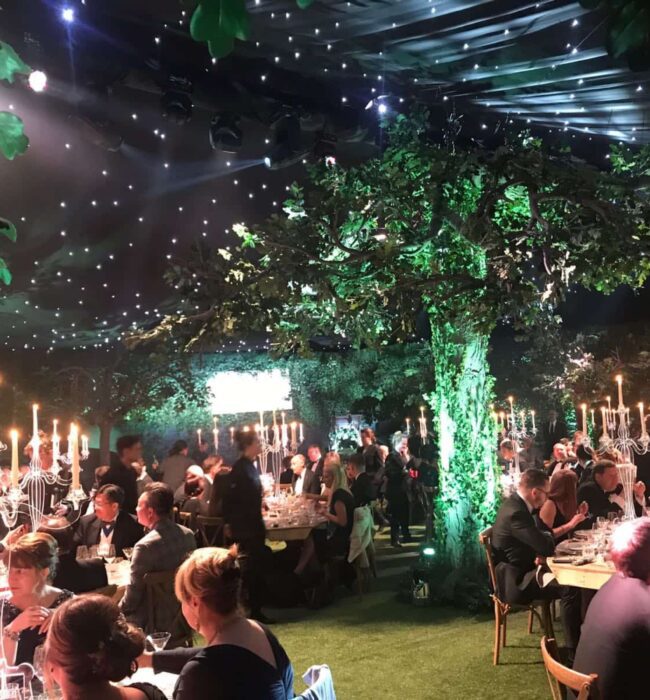 Large artificial tree for hire, uplit green in a candlelit marquee with starry ceiling lights and diners at tables