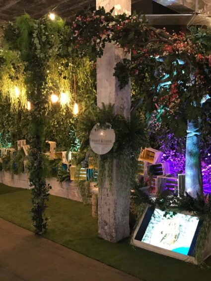 Artificial ivy-wrapped pillars and blossom tree for hire, on an exhibition stand with crates, bulb lights and grass