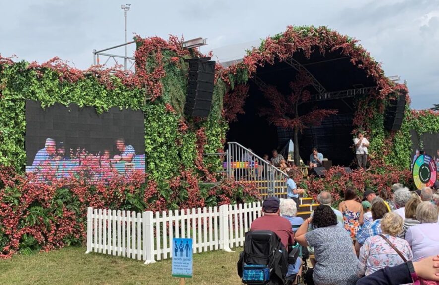 Artificial ivy and red blossom walls for hire, framing a festival stage with a screen and white picket fence