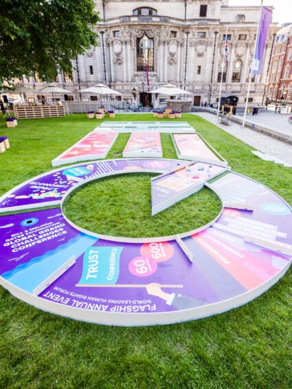 Large circular crazy golf course with printed greens on a lawn outside the QEII Centre, with wooden box seats