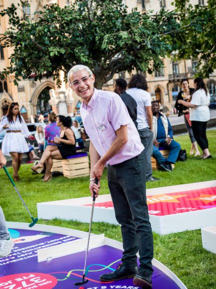 Man putting on a crazy golf course on a lawn at the QEII Centre, with spectators on wooden box seats behind