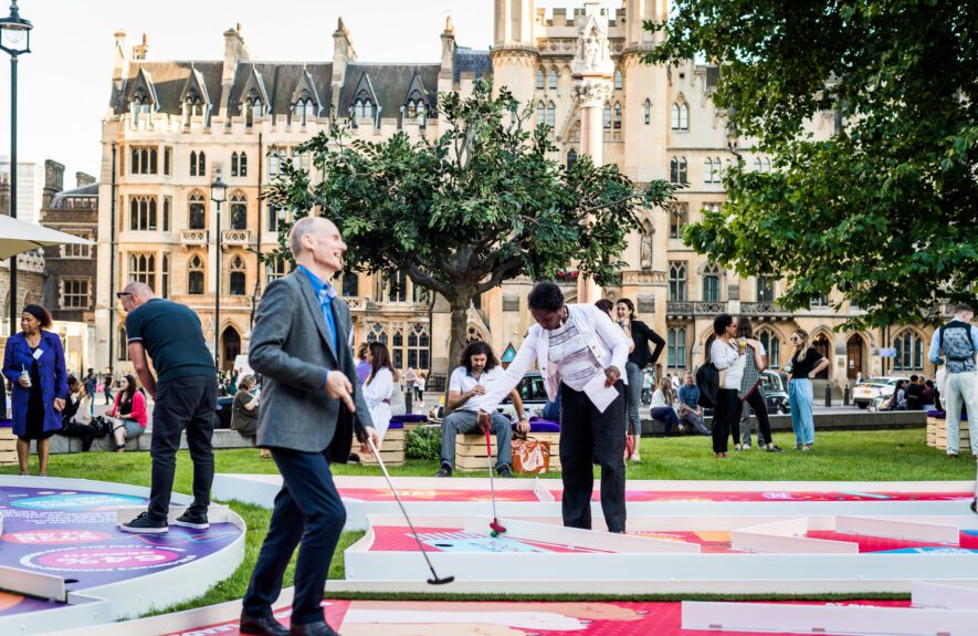 Guests playing crazy golf on a lawn in front of a Gothic building, with spectators on wooden box seats