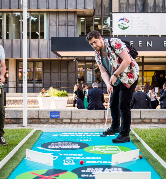 Man in a floral shirt putting on a blue crazy golf hole printed 'Minimal Waste' outside the QEII Centre