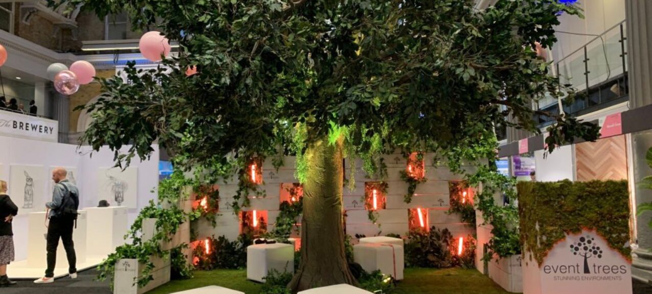 Large artificial tree for hire, placed on artificial grass with white cube seats and a branded plinth at an exhibition