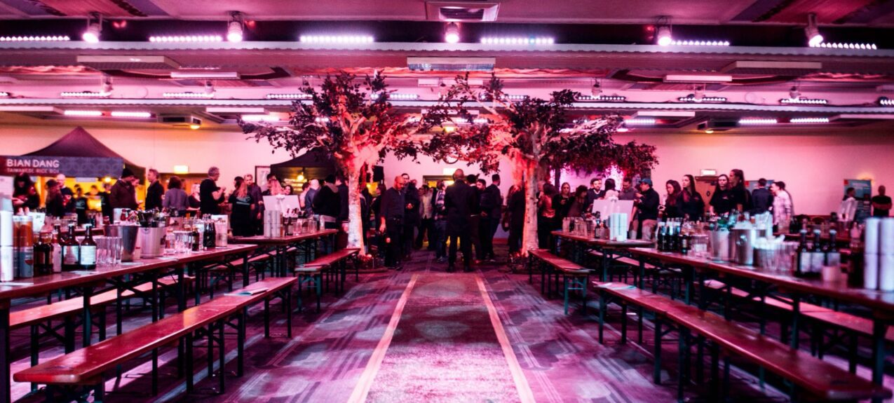 Artificial leafy trees for hire at the end of a hall of long bench tables, lit pink, with guests and bars