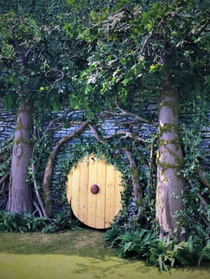 Round wooden door in a stone wall framed by two artificial trees, ivy and foliage, for a themed garden set