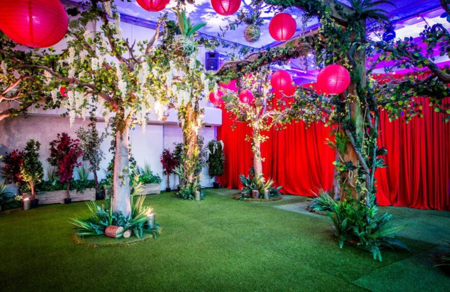 Artificial trees with trailing white wisteria for hire on artificial grass, with red paper lanterns and red curtains
