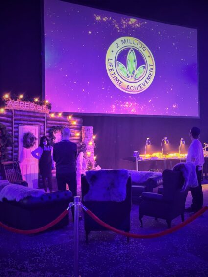 Log cabin apres ski set with fur armchairs, wreaths and fairy lights, beside a 2 MILLION LIFETIME ACHIEVEMENT screen