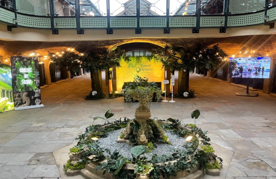 Artificial foliage and plants for hire around a courtyard fountain, with a CHECK IN HERE sign and Event Odyssey backdrop