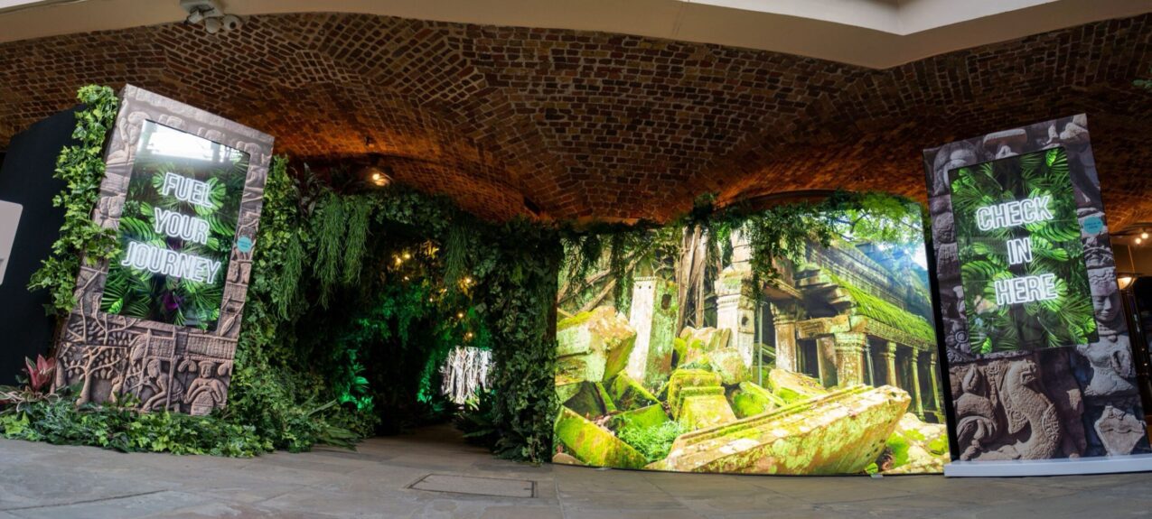 Artificial jungle foliage wall for hire at a brick-vaulted entrance, with FUEL YOUR JOURNEY and CHECK IN HERE screens