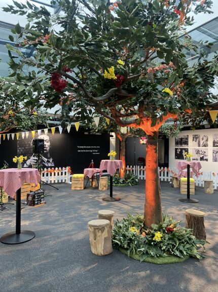 Artificial tree with orange-lit trunk and coloured flowers for hire, with bunting and picket fencing indoors