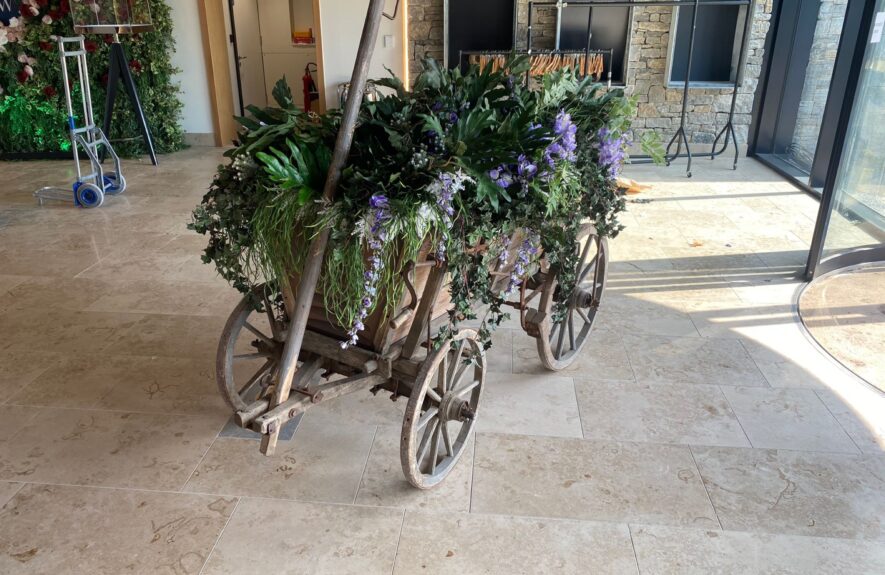 Old wooden cart filled with artificial foliage and trailing purple wisteria for hire, in a stone-floored venue lobby