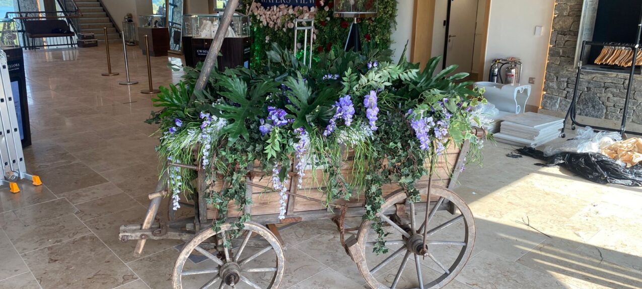 Wooden cart planted with artificial foliage and purple wisteria for hire, in front of a rose and foliage wall, Waters View