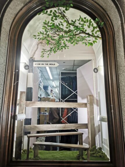 Artificial leafy tree branch for hire, in an arched Regent Street window with a 'Stow on the Wold' signpost and wooden stile