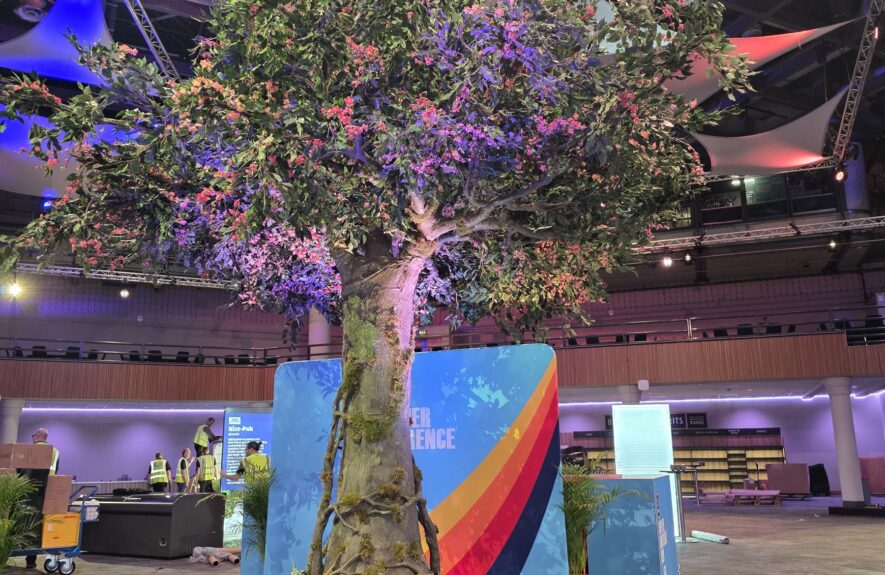 Large artificial blossom tree for hire, on a mossy base in front of a rainbow-striped exhibition stand at a conference