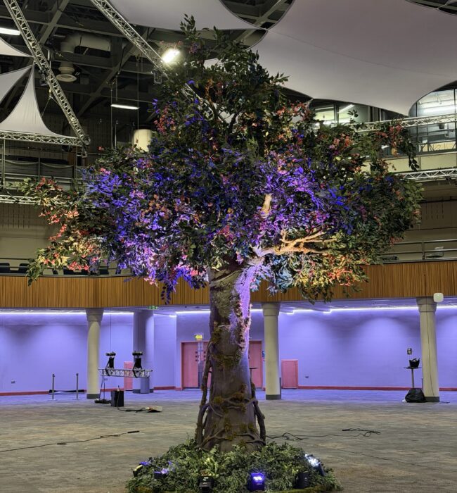 Artificial blossom tree for hire, with purple uplighting at its base and white ceiling sails in a conference hall