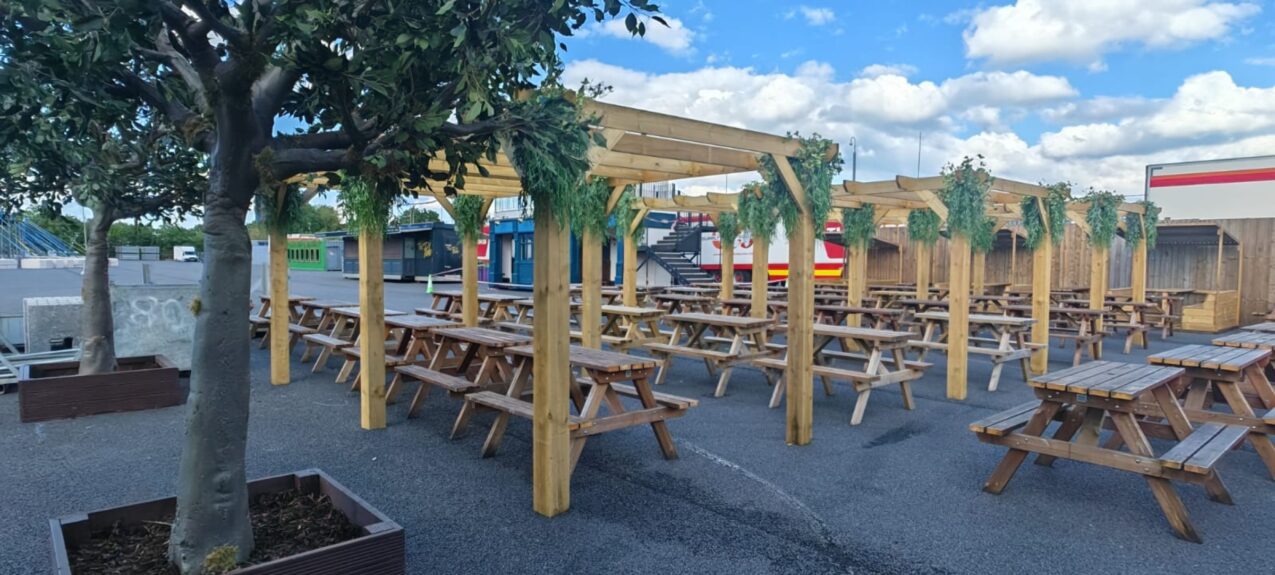 Artificial tree for hire, in a timber planter beside wooden pergolas with picnic benches at Download Festival