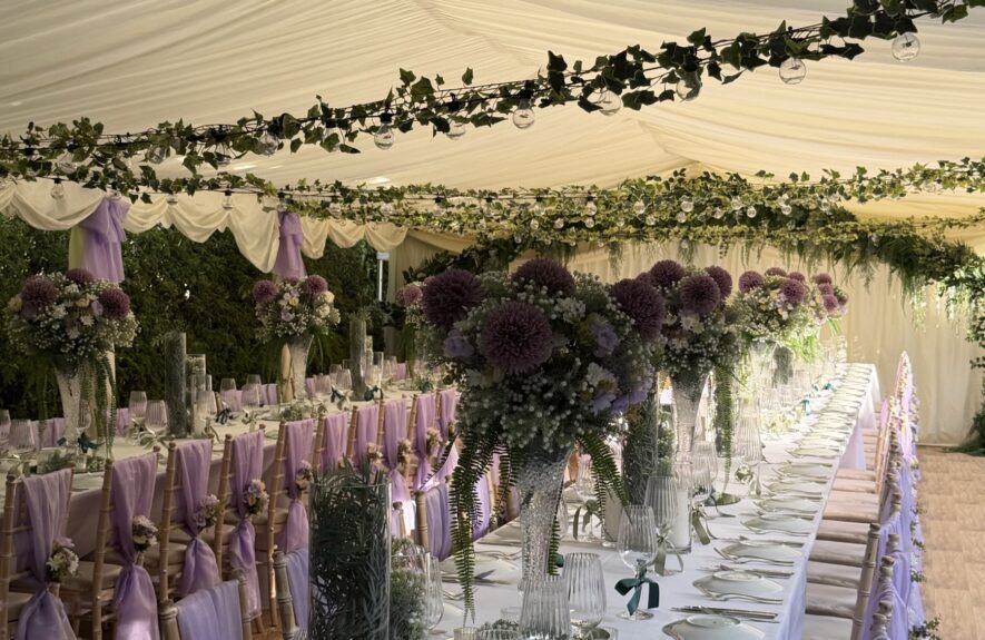 Artificial ivy garlands for hire, hung over long marquee wedding tables, with festoon bulbs and purple pompom flowers