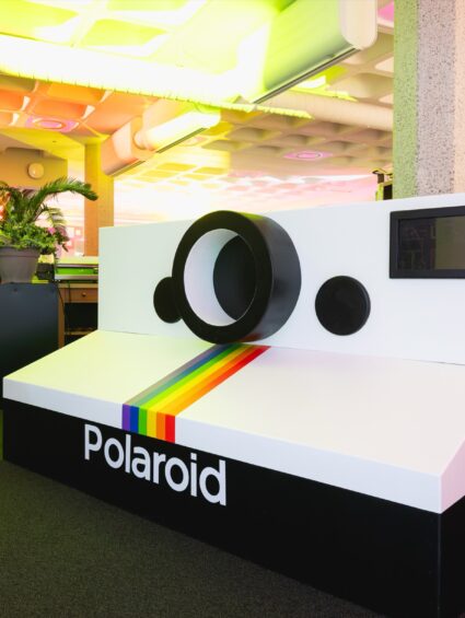 Giant Polaroid camera prop at the QEII Centre 40th anniversary showcase, with a plant in a black planter behind